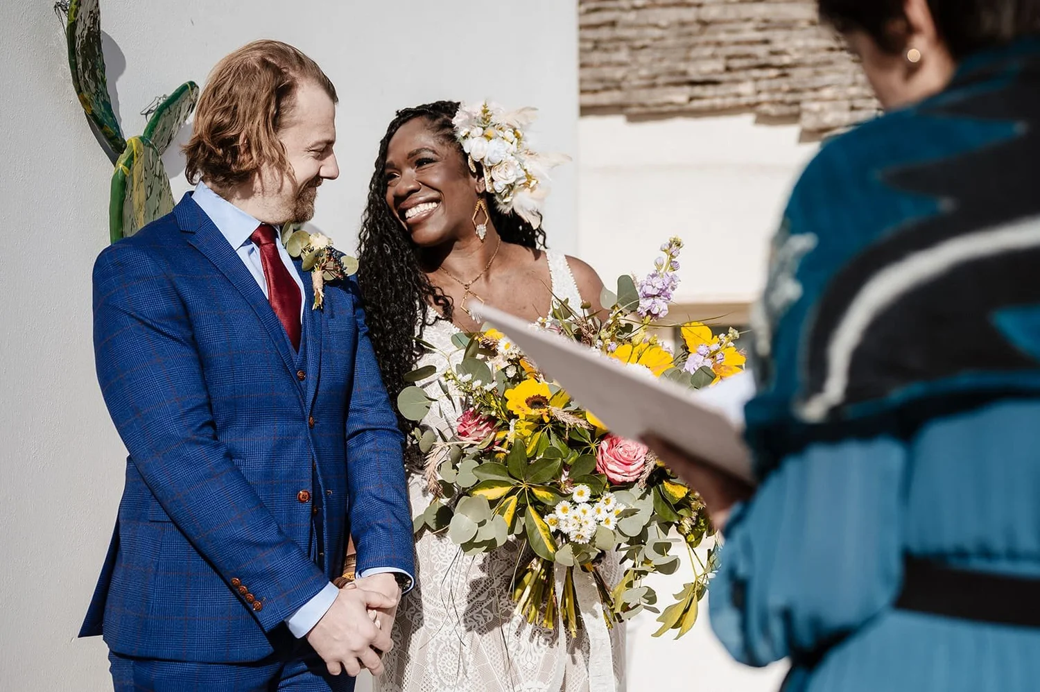 A same-sex couple getting married, smiling and holding hands, with an officiant reading vows. The bride wears floral accessories and holds a large bouquet, while the groom wears a blue suit. The scene takes place indoors with a colorful wall decor in