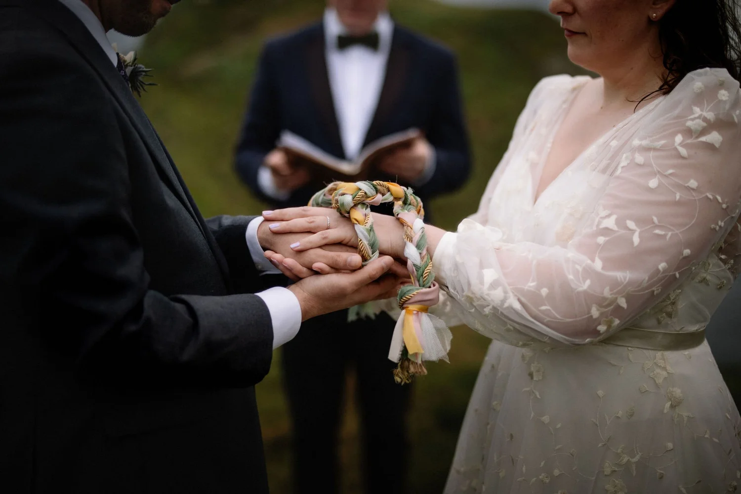 A couple exchanging wedding vows, holding hands with a decorative ribbon, with an officiant in the background.