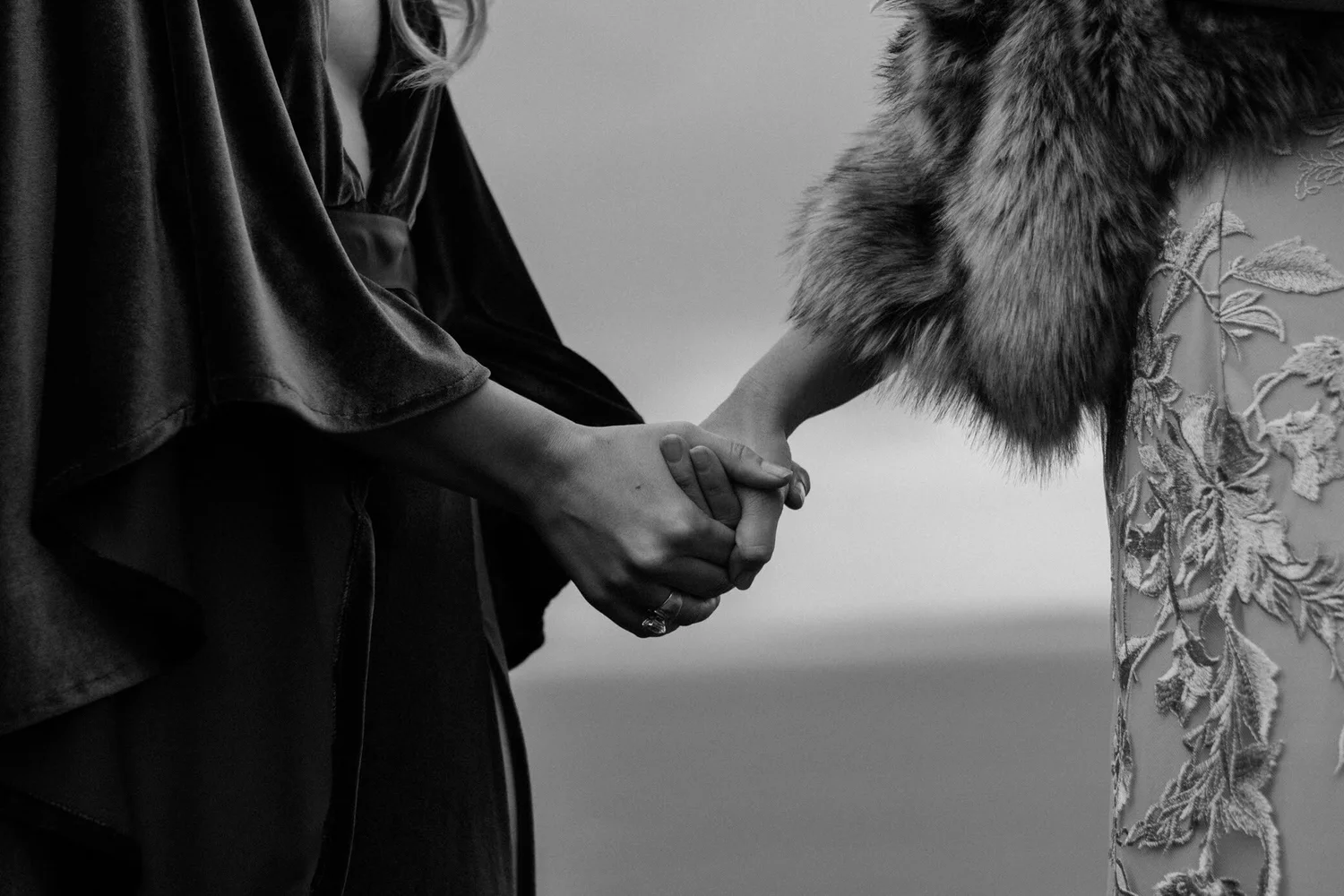 A close-up black and white photo of two people holding hands, one in a dark leather jacket and the other in a dress with embroidered floral details.