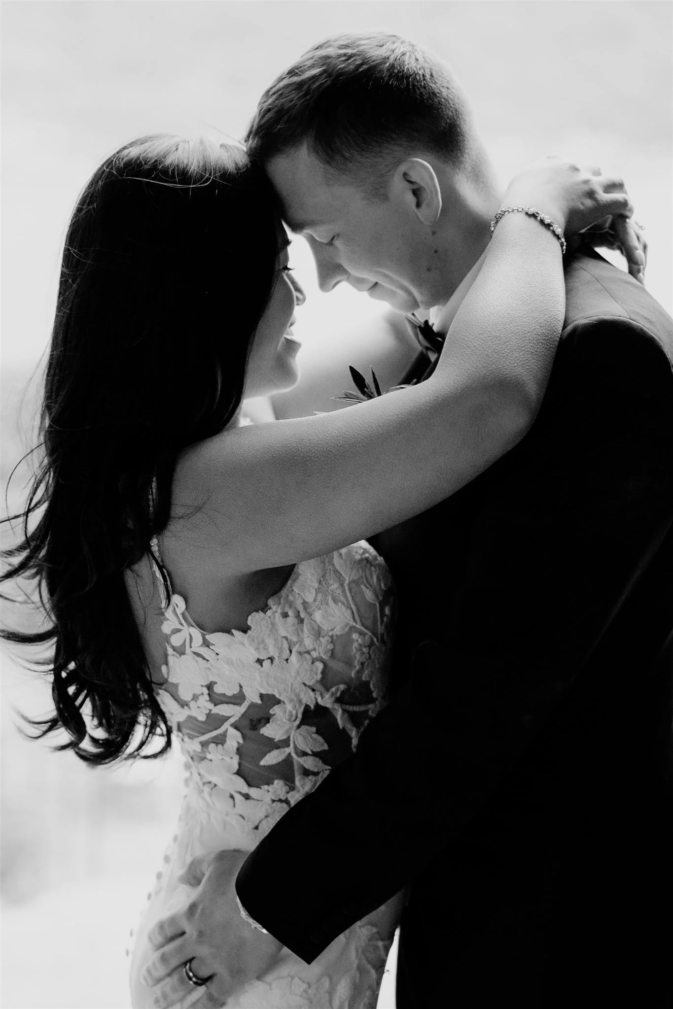 A black and white photo of a couple embracing, with their foreheads touching and eyes closed. The woman is wearing a lace wedding dress, and the man is wearing a suit. The woman's arm is around the man's neck, and the man's hand is resting on the wom