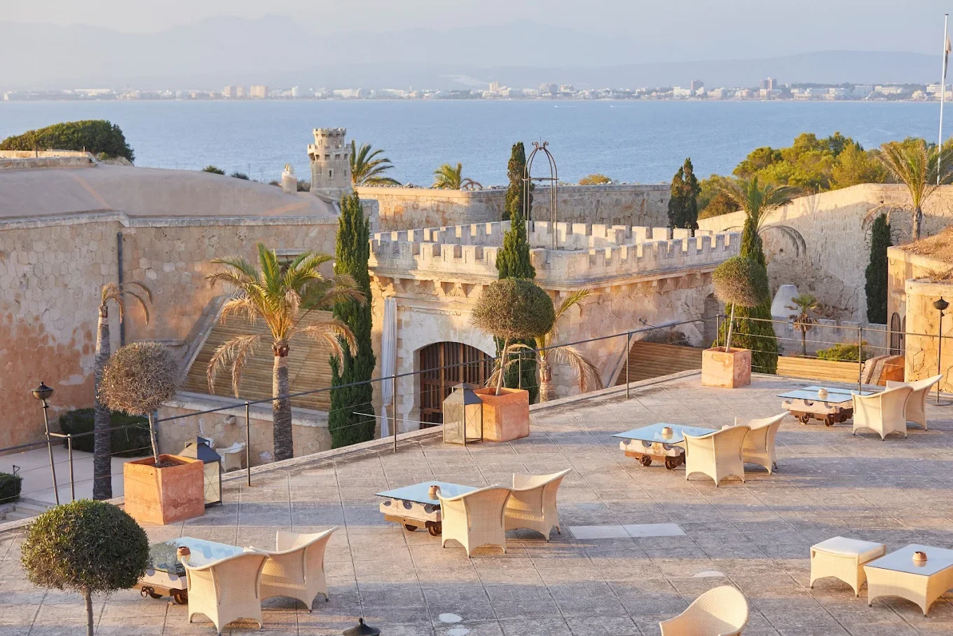 Terrace with tables and chairs overlooking historic stone buildings, trees, and waterfront in distance.