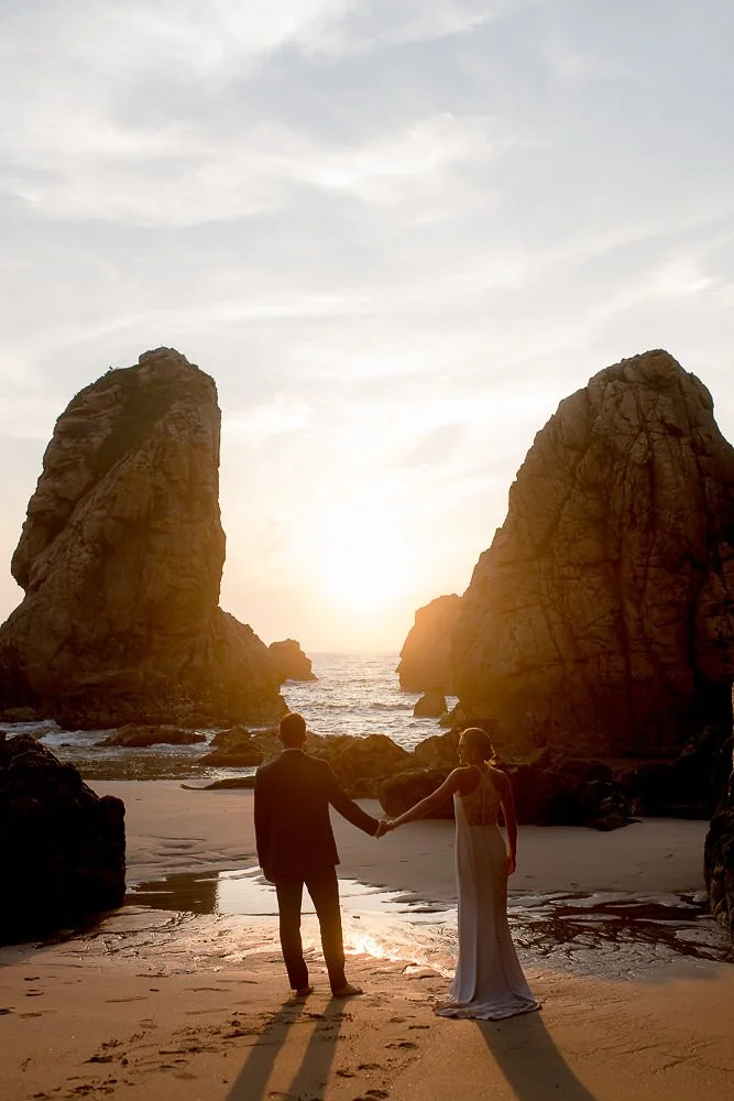A couple holding hands on a beach at sunset, with large rock formations and the ocean in the background.