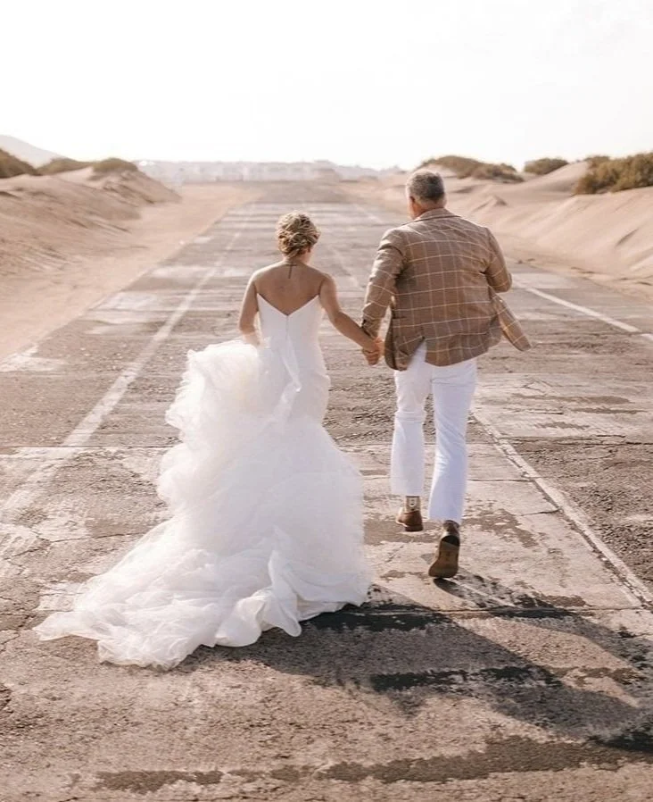 Bride and groom walking hand-in-hand on an empty desert road, dressed in wedding attire, with sandy dunes on either side.