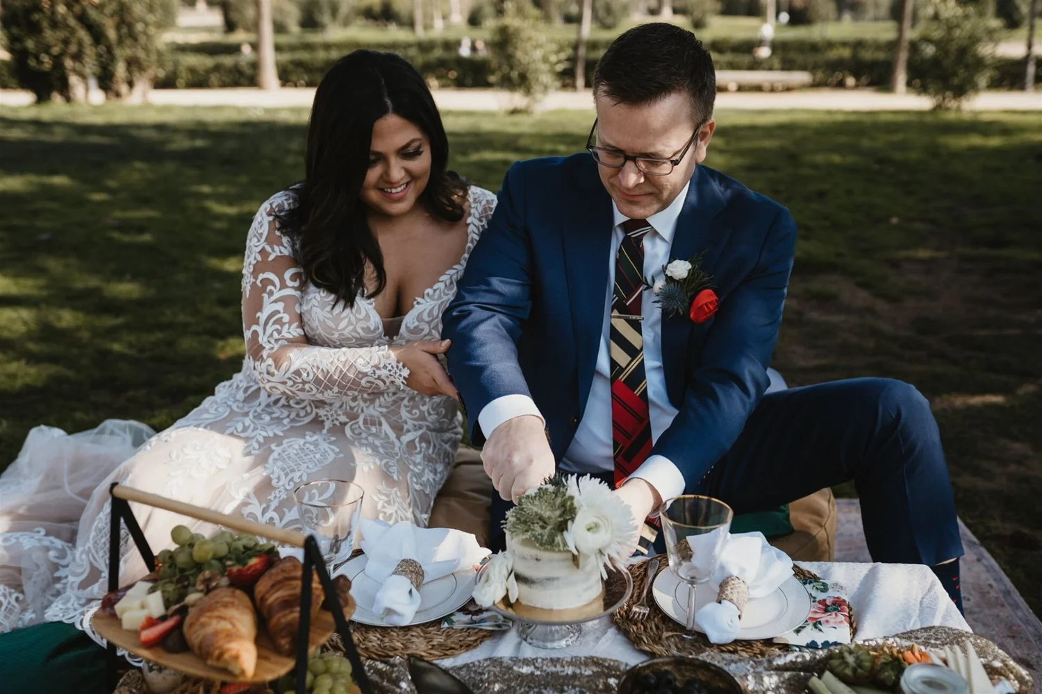 A newlywed couple at a wedding celebration outdoors, sitting at a table with food and drinks, sharing a moment of happiness.