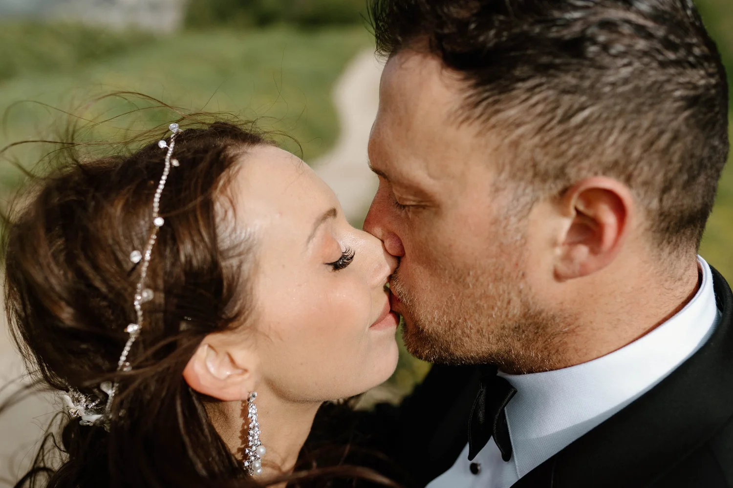 A close-up of a man and woman with their eyes closed, noses touching, sharing a kiss outdoors, dressed in formal attire, with the woman wearing jewelry and a decorative hairpiece.
