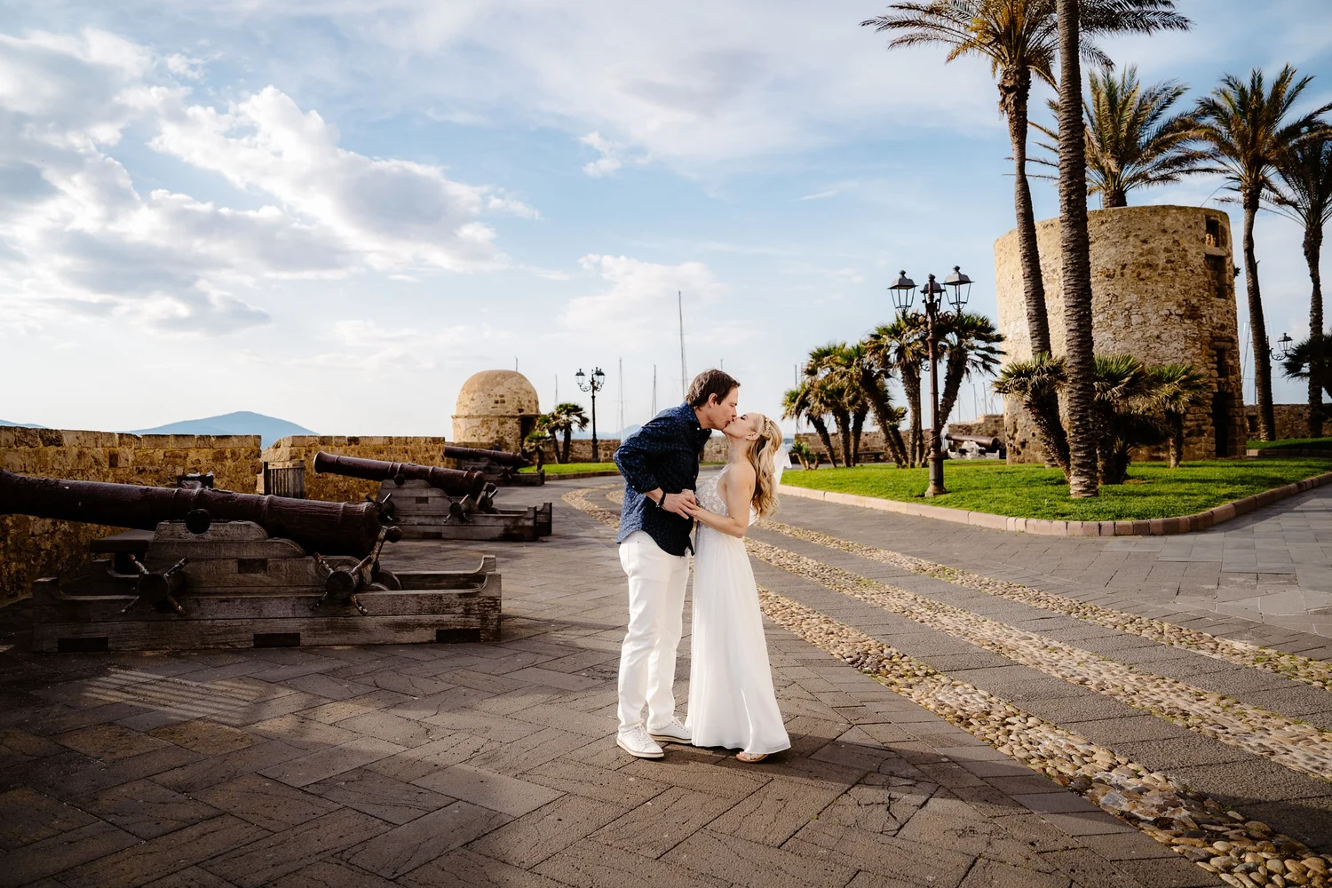 A couple dressed in wedding attire sharing a kiss outdoors near historic stone walls, palm trees, and old-fashioned street lamps under a partly cloudy sky.