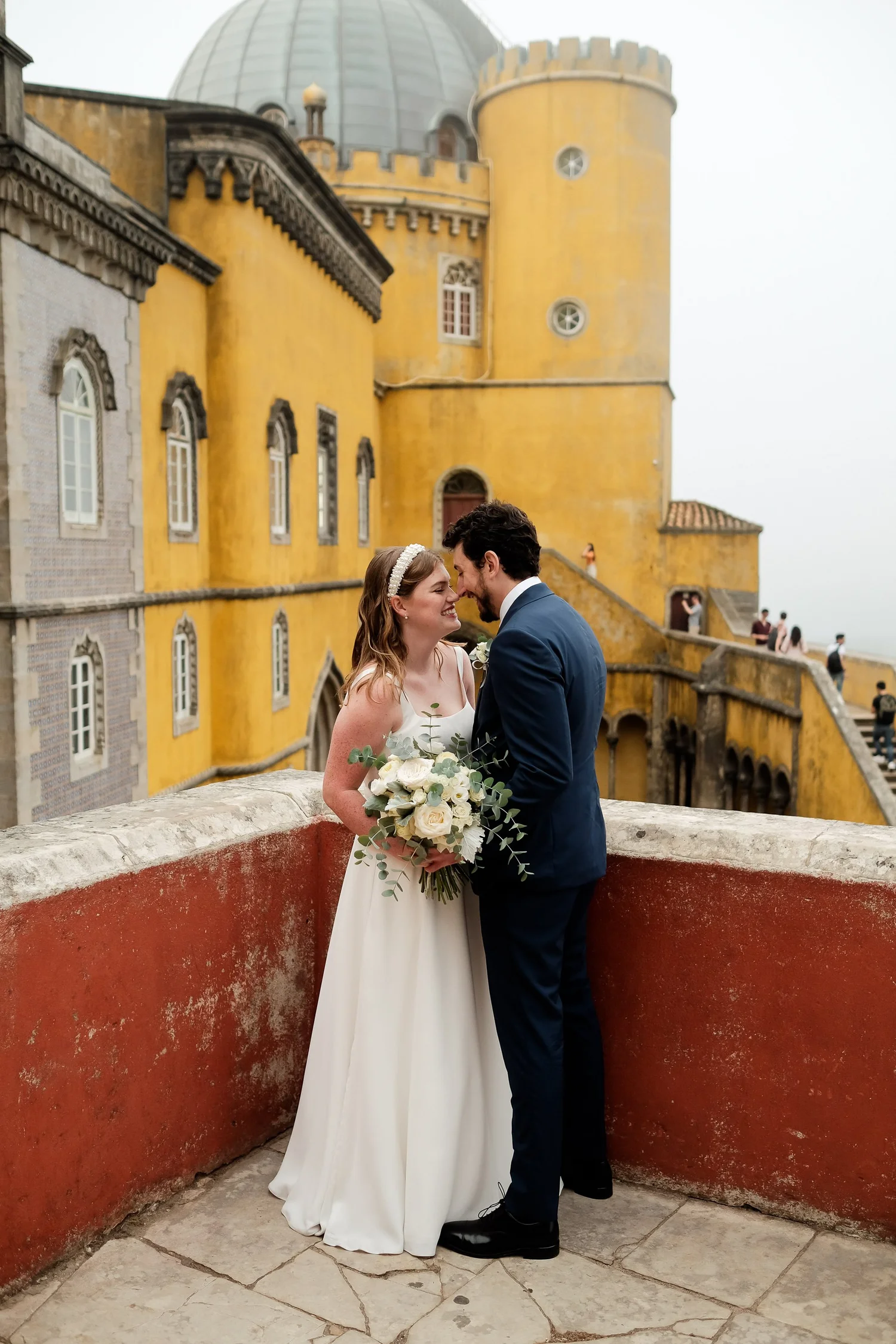 A bride and groom are standing close together on a terrace overlooking a yellow castle-like building. The bride is holding a bouquet of white roses and greenery, and the groom is wearing a navy blue suit. They are smiling at each other with their foreheads touching.