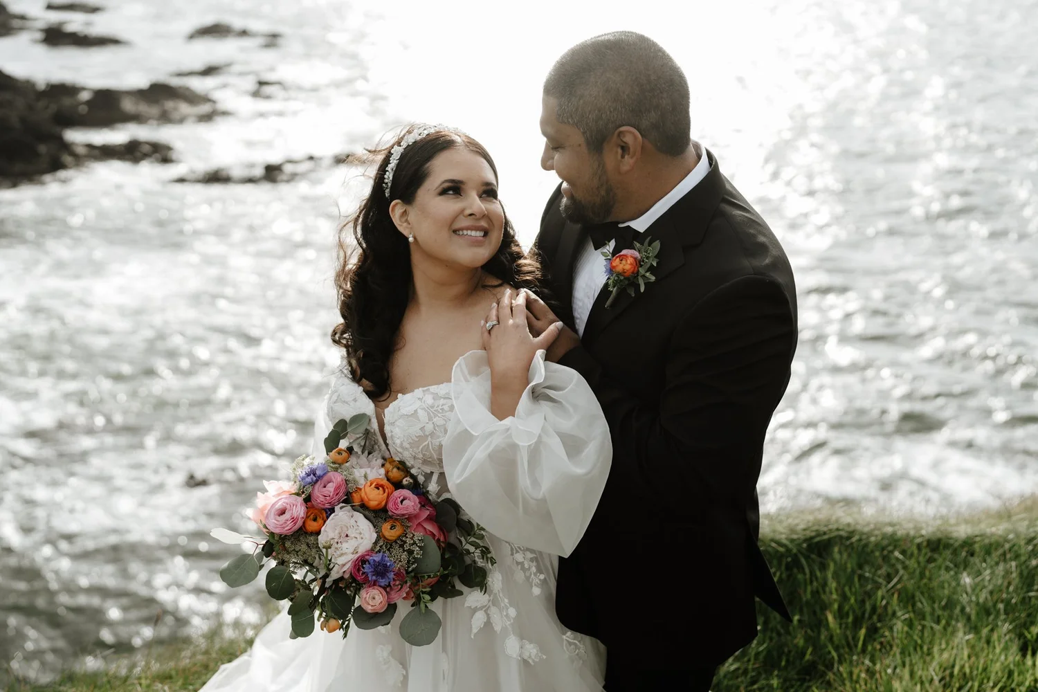 A bride and groom sharing a romantic moment near a body of water, with the bride holding a colorful bouquet of flowers, dressed in a white wedding gown and the groom in a black tuxedo with a boutonnière.