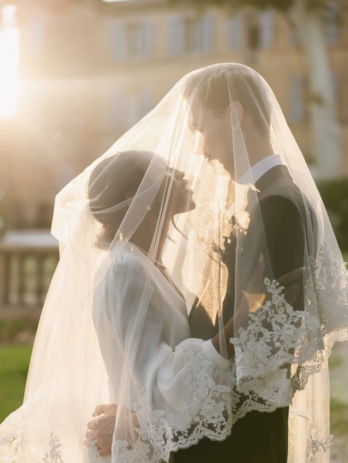 A bride and groom sharing a kiss under a wedding veil outdoor, with sunlight and trees in the background.