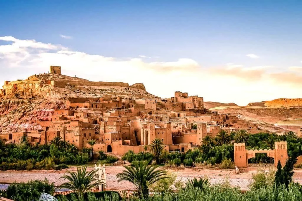 Pics of a desert castle built on a hillside with painted brown walls and palm trees in the foreground.