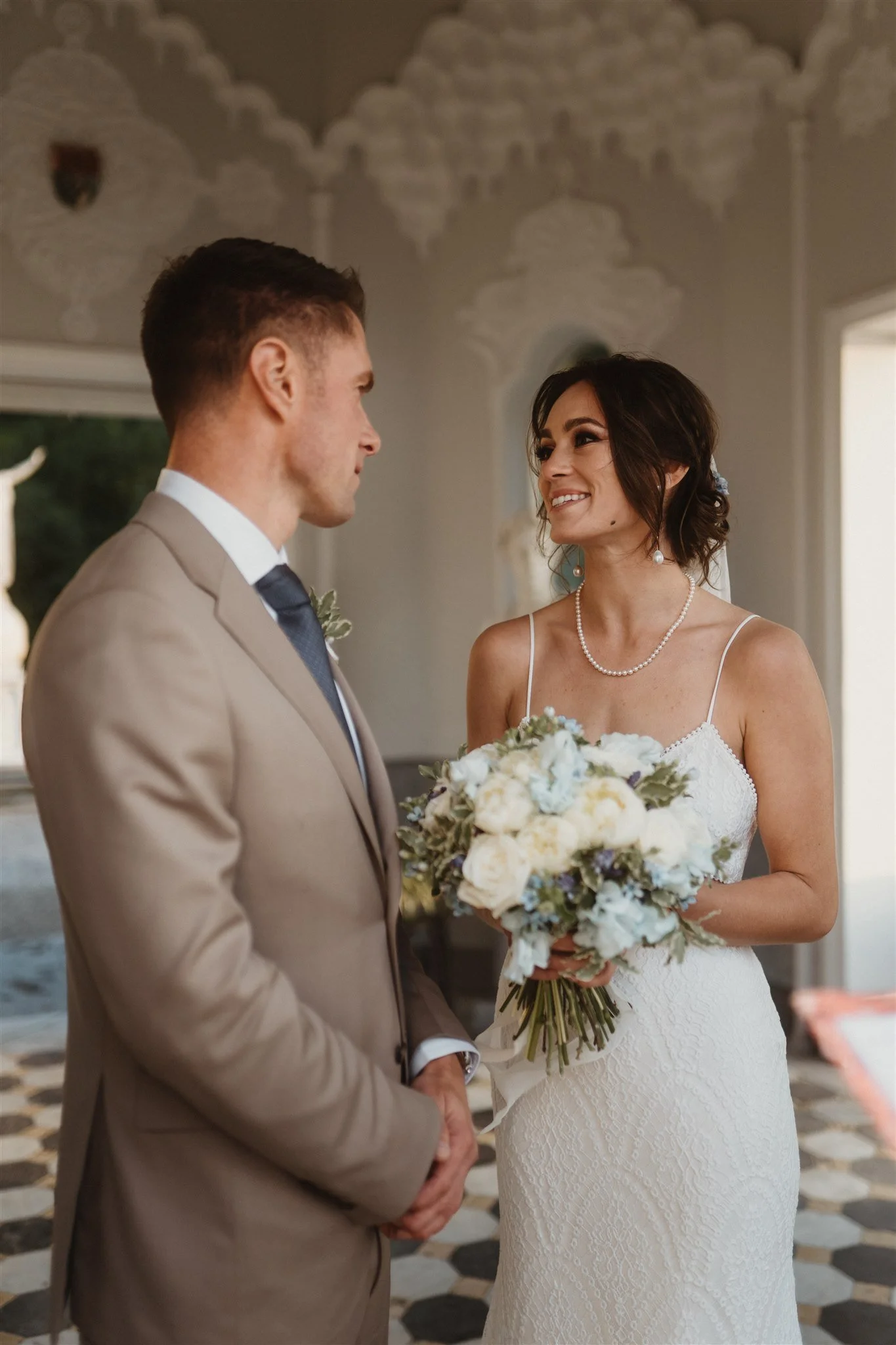 A bride and groom facing each other during a wedding ceremony, with the bride holding a bouquet of white and purple flowers, both smiling softly.