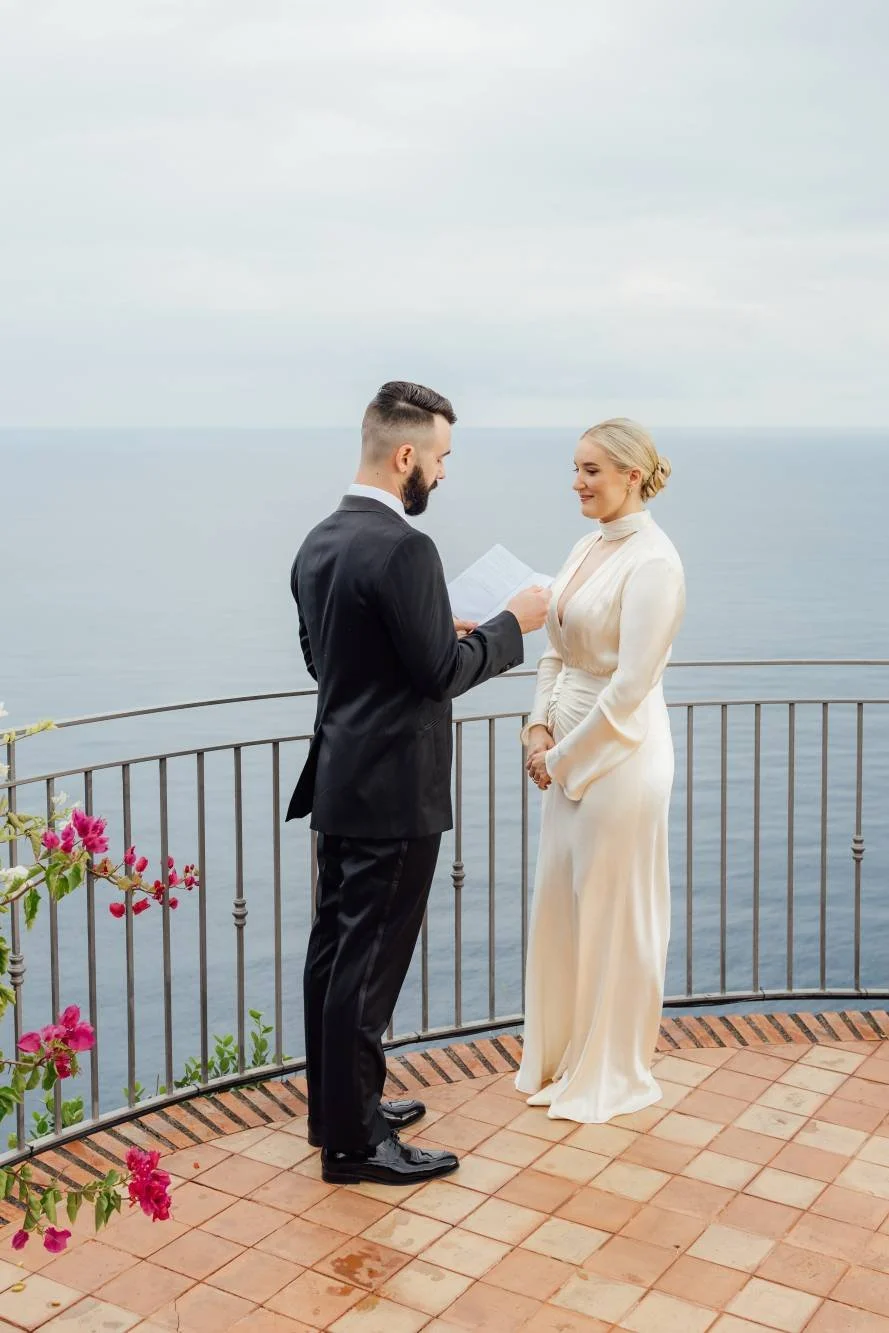 A couple getting married on a balcony overlooking the ocean, with the groom reading vows to the bride.