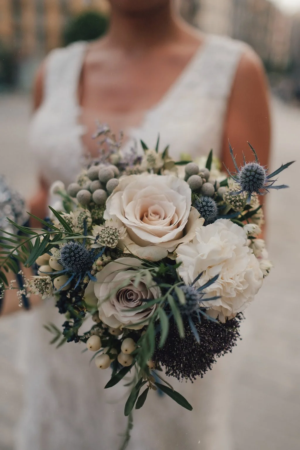 A bride holding a bouquet of white roses, hydrangeas, and blue thistles, with greenery, against a blurred outdoor background.