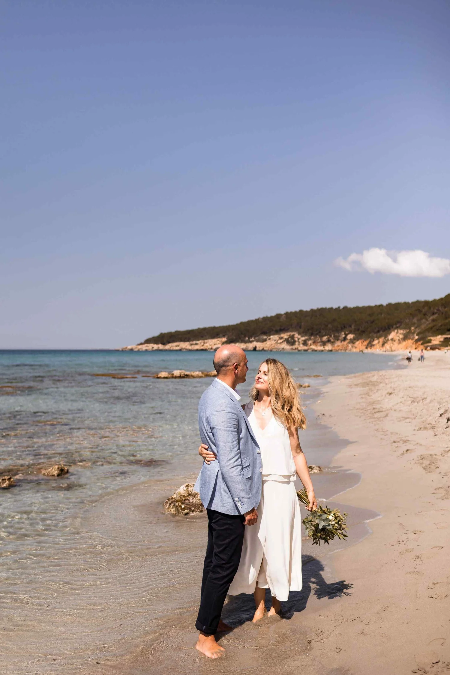 A couple stands close together on a beach with gentle waves and rocky shoreline, embracing and looking into each other's eyes during a sunny day, with a clear blue sky and land in the background.