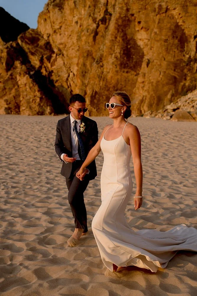 A smiling woman in a white wedding dress and sunglasses holding hands with a man in a dark suit and tie on a sandy beach with large rocky cliffs in the background.
