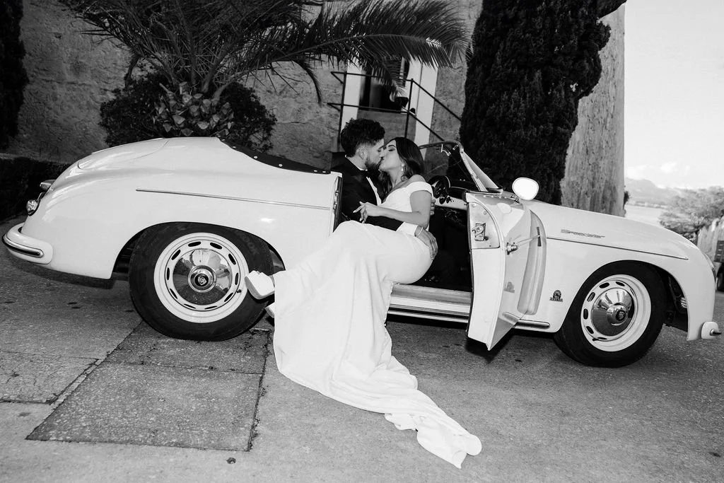 A couple sits on the edge of a vintage convertible car, sharing a kiss. The scene is outdoors near a stone wall, trees, and a building, in black and white.