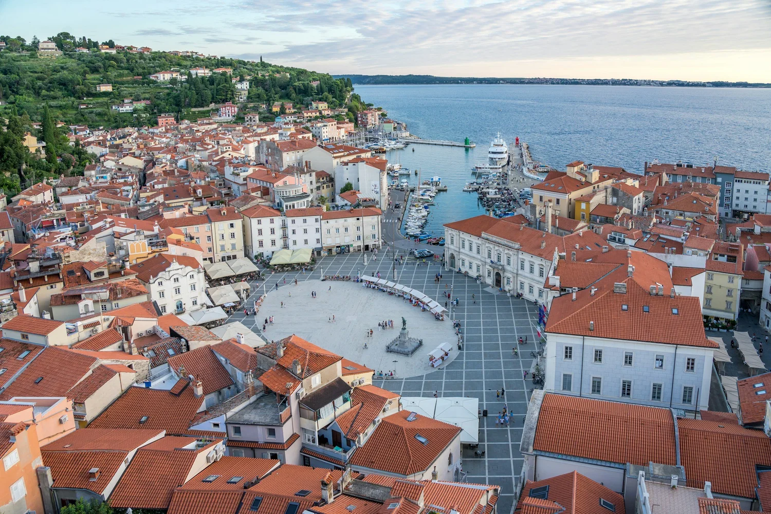 Aerial view of a coastal town with red-tiled roofs, a small harbor with boats, a large open square with people, and lush green hills in the background.