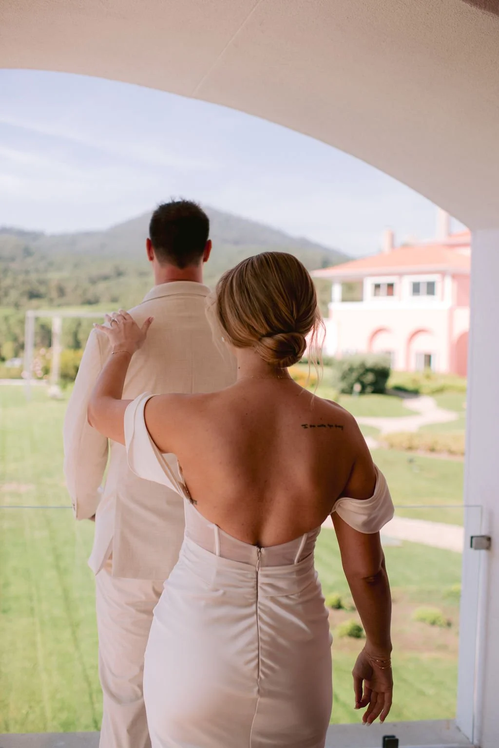 A woman with blonde hair in a bun, wearing a white off-shoulder dress, gently touching a man in a cream-colored suit, with a scenic outdoor view of greenery and a pink building in the background.