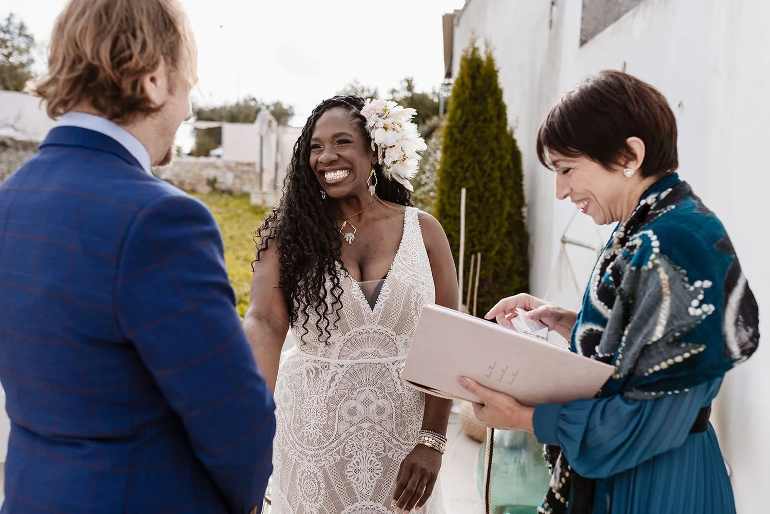 A woman in a white lace dress with a floral headpiece smiling as she is getting married, standing outside with a man and another woman who is holding a binder, both smiling.