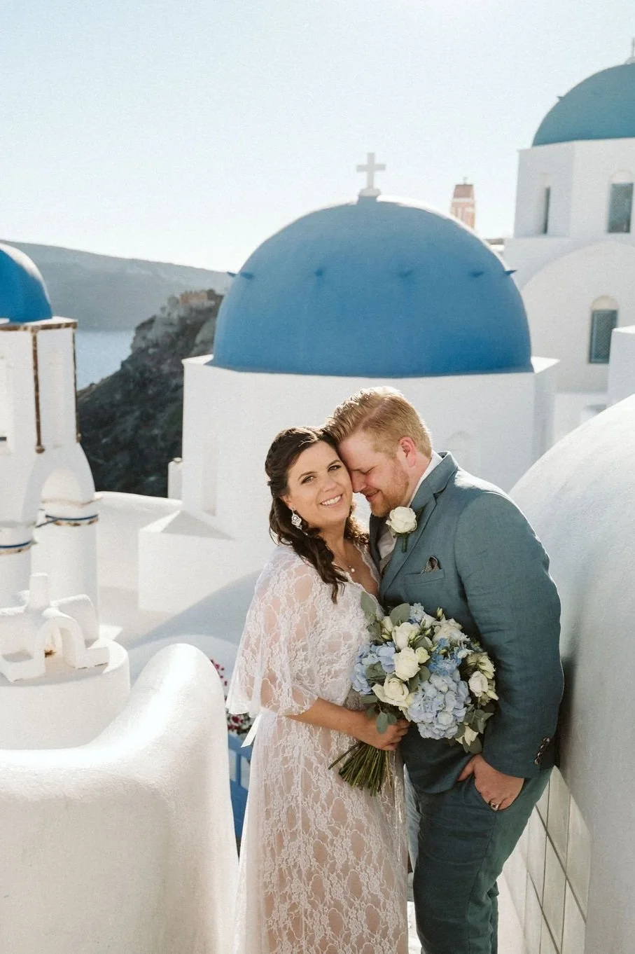 A newlywed couple smiling and embracing on a rooftop terrace with white buildings and blue domes in Santorini, Greece, during daytime.