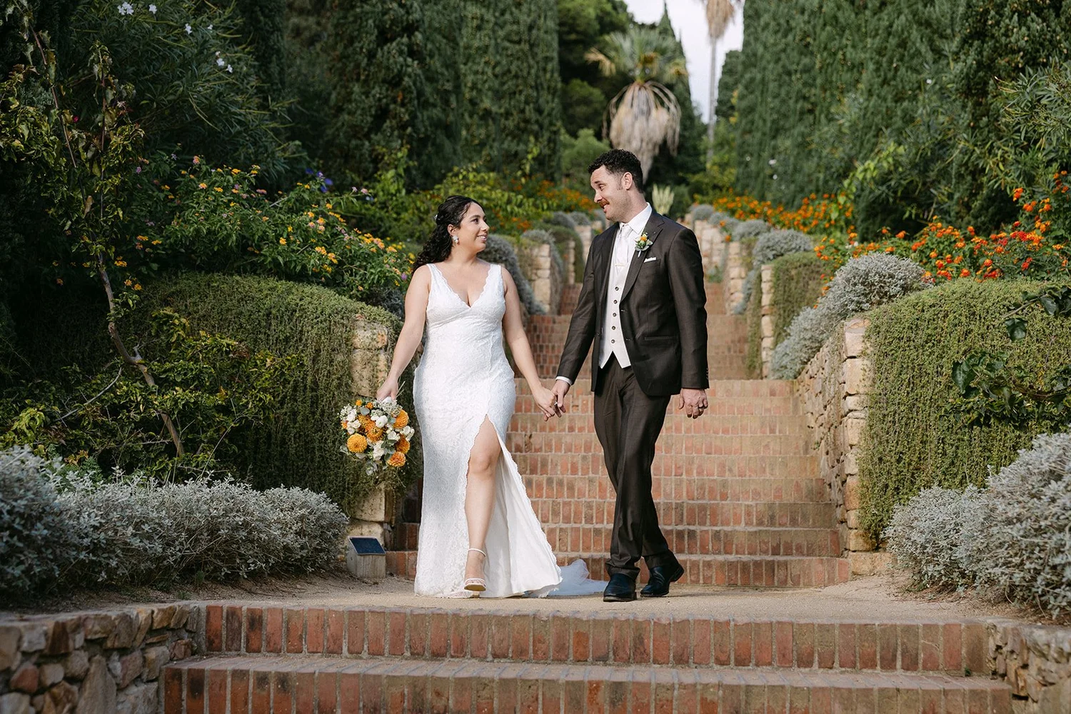 A bride and groom holding hands and walking down brick steps in a garden. The bride is wearing a white lace wedding gown and holding a bouquet of flowers. The groom is dressed in a dark suit with a boutonniere. They are smiling at each other amidst l