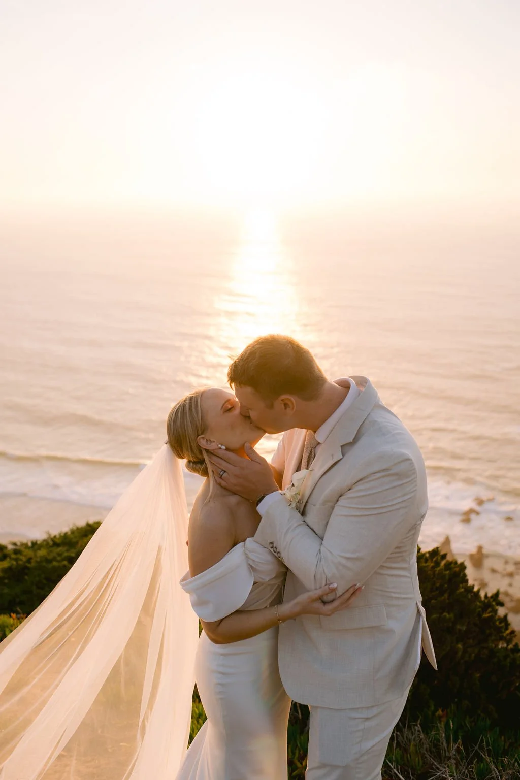 A bride and groom sharing a kiss during sunset at the beach, with the ocean and sunset in the background.