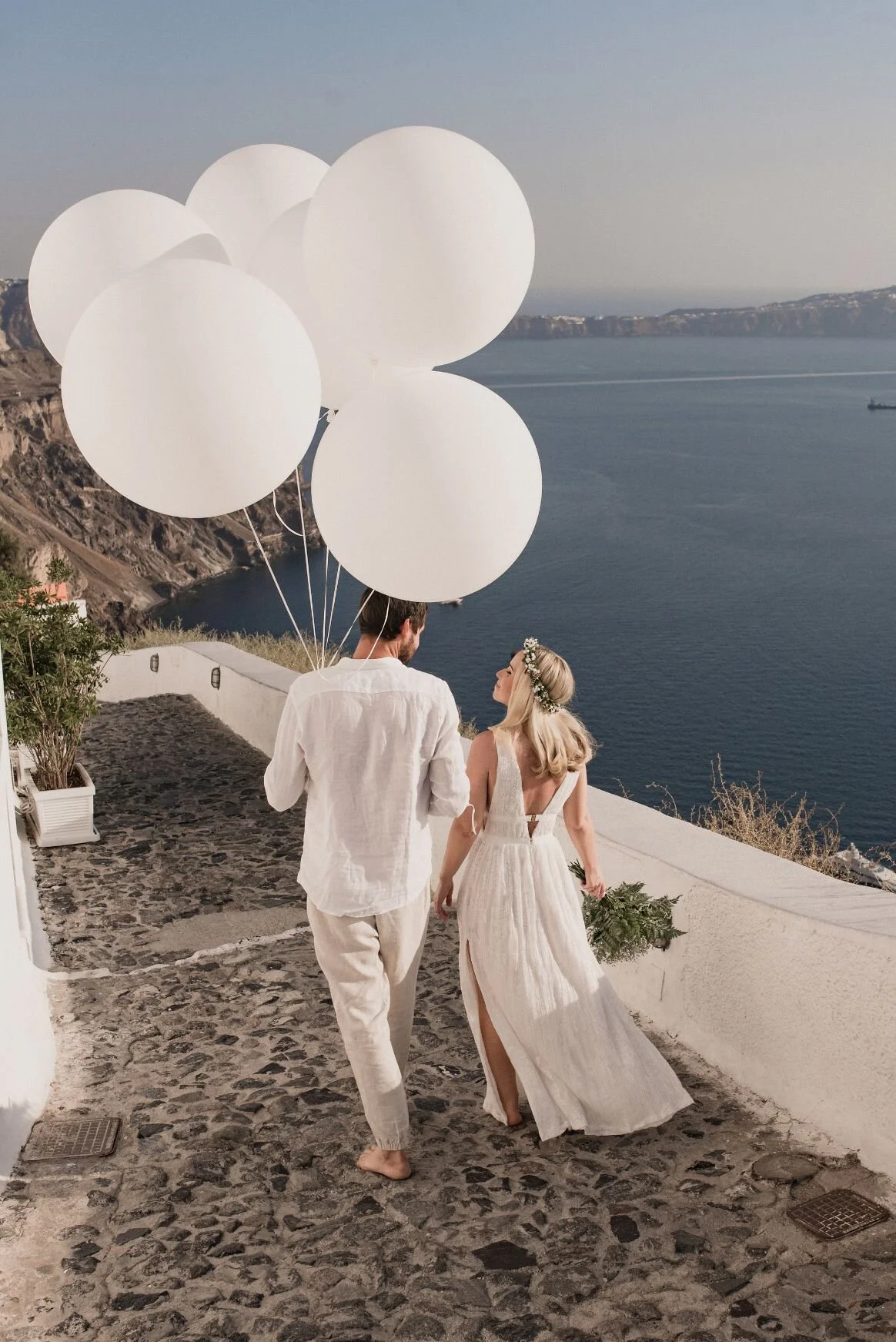 A couple dressed in white walking along a cobblestone pathway by the sea, holding white balloons and a bouquet, with a scenic view of the water and coastline in the background.