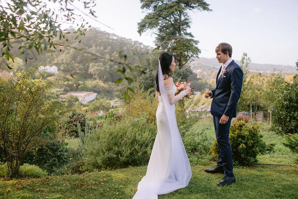 A bride and groom stand outdoors in a garden, facing each other. The bride wears a white wedding gown and veil, holding a bouquet of flowers. The groom wears a navy suit and white shirt, holding a glass of wine. The background features trees, bushes, and a distant hilly landscape under a partly cloudy sky.