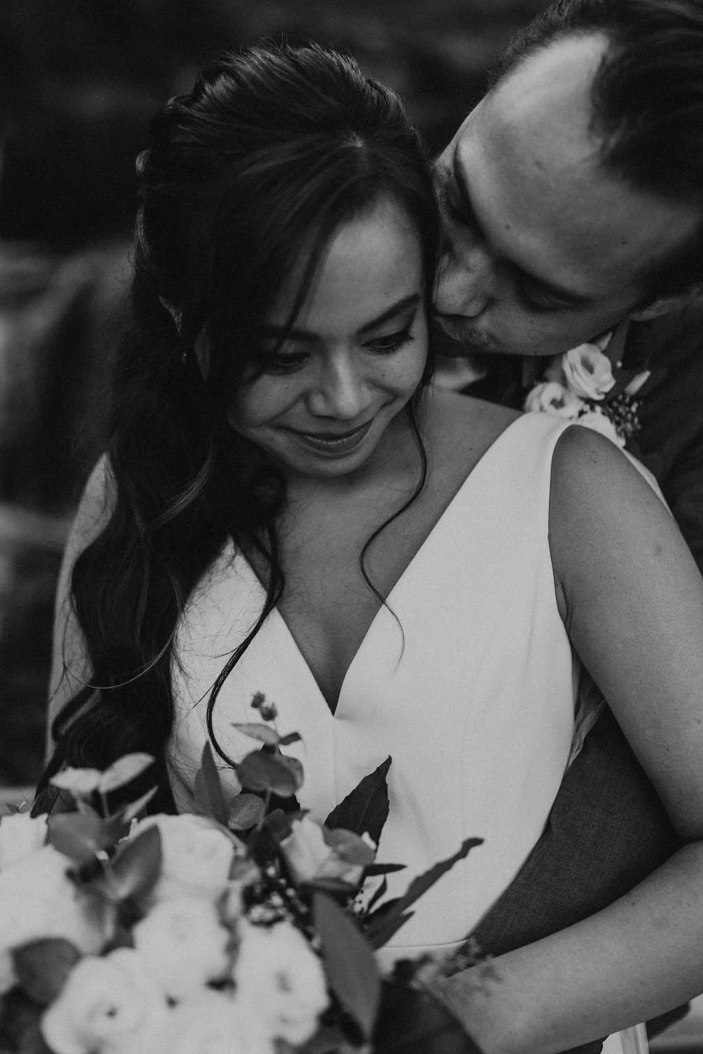 Black and white photo of a couple embracing closely, with the man kissing the woman's cheek and the woman smiling down. The woman has long wavy hair and is wearing a sleeveless dress, holding a bouquet of flowers.