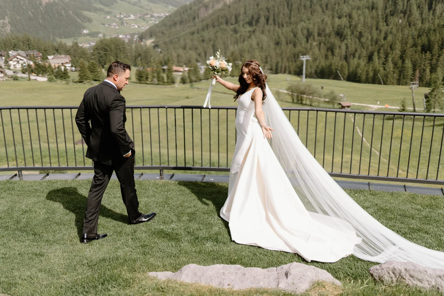A bride in a white wedding dress and veil is smiling and holding a bouquet of flowers as she dances with a groom in a black suit and tie on a grassy area in front of a scenic mountain landscape.