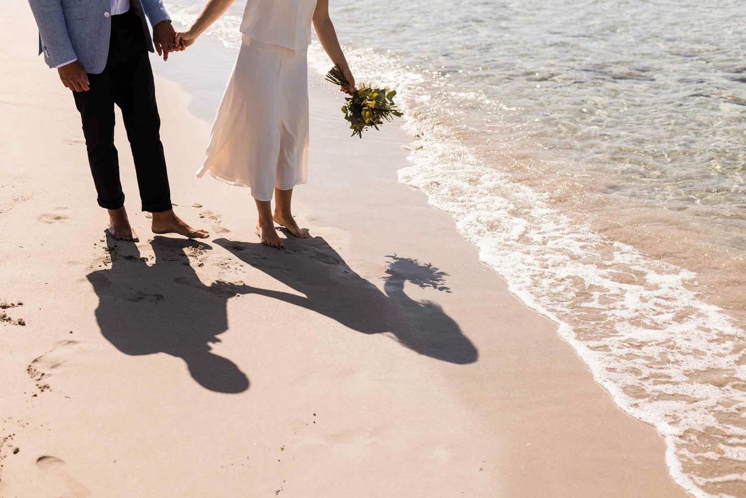 A couple holding hands walking along the beach shoreline with their shadows cast on the sand, nearby a bouquet of flowers in the woman's hand.