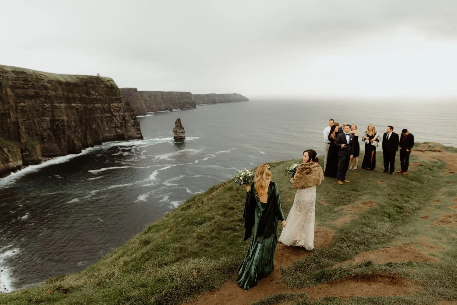 A bride and groom standing on a grassy cliff overlooking the ocean, with wedding guests nearby, during an overcast day.