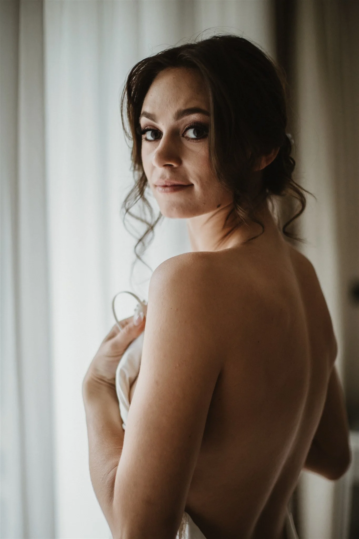 A woman with dark, wavy hair looking over her shoulder near a sheer curtain.