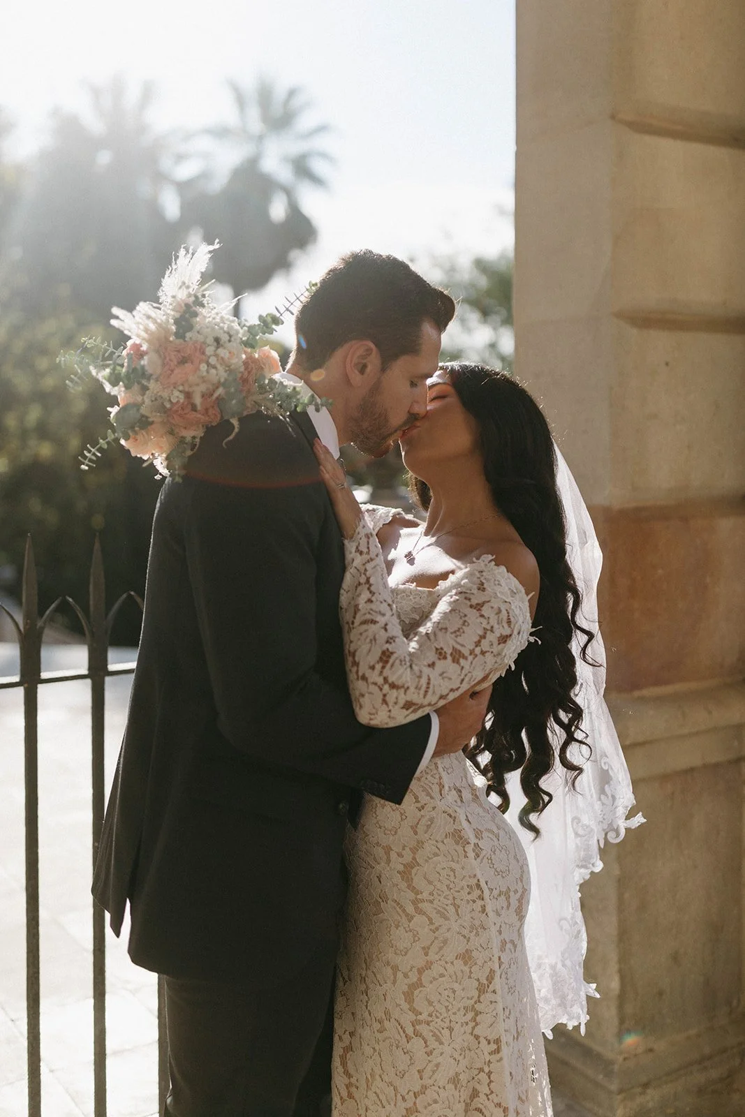 A couple sharing a kiss on their wedding day, with the bride wearing a lace dress and veil, and the groom in a black suit, outdoors during daytime, with palm trees in the background.