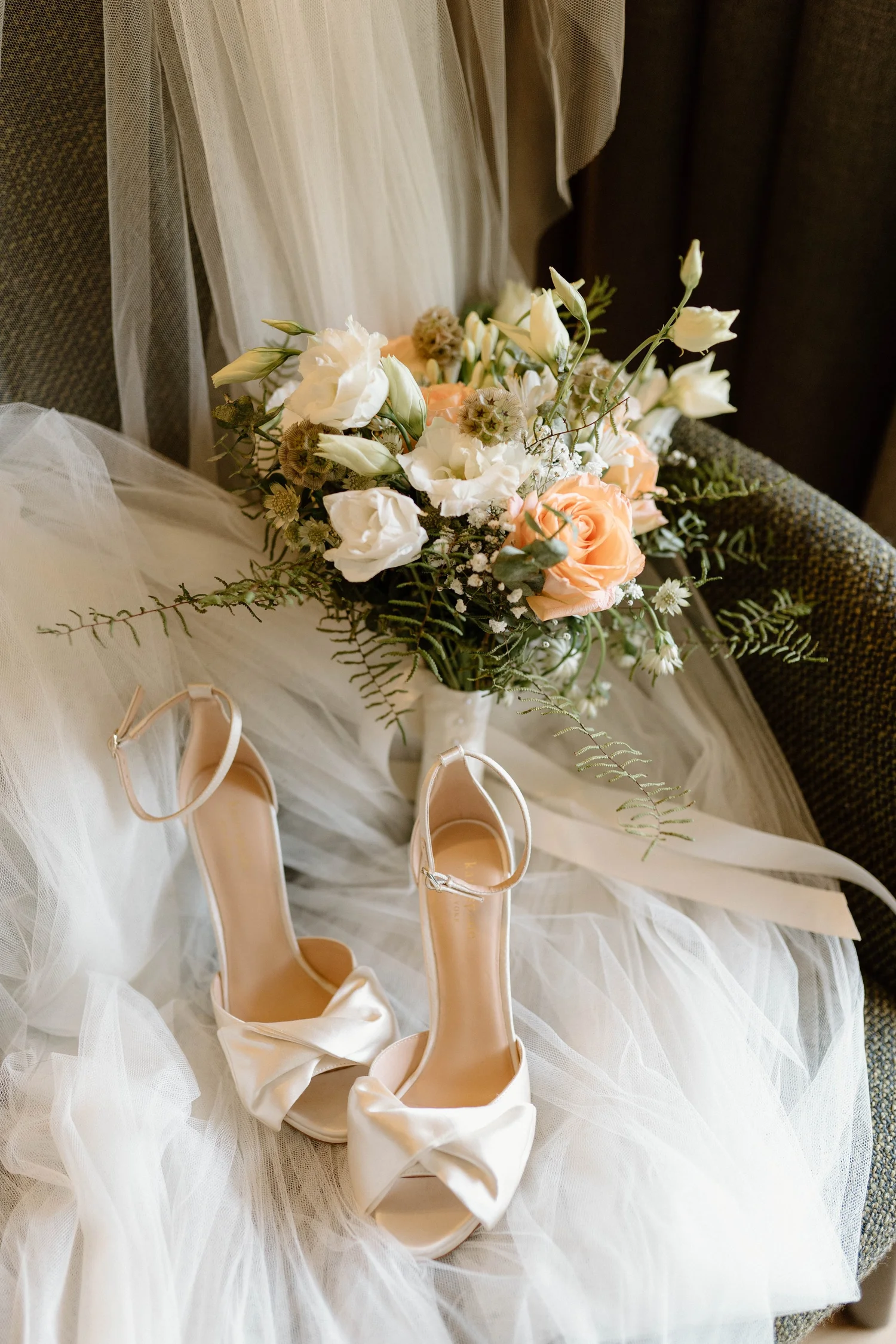 A pair of cream-colored satin bridal shoes with bows, a floral bouquet with white and peach roses, white lisianthus, and greenery, resting on a white, tulle-covered surface.