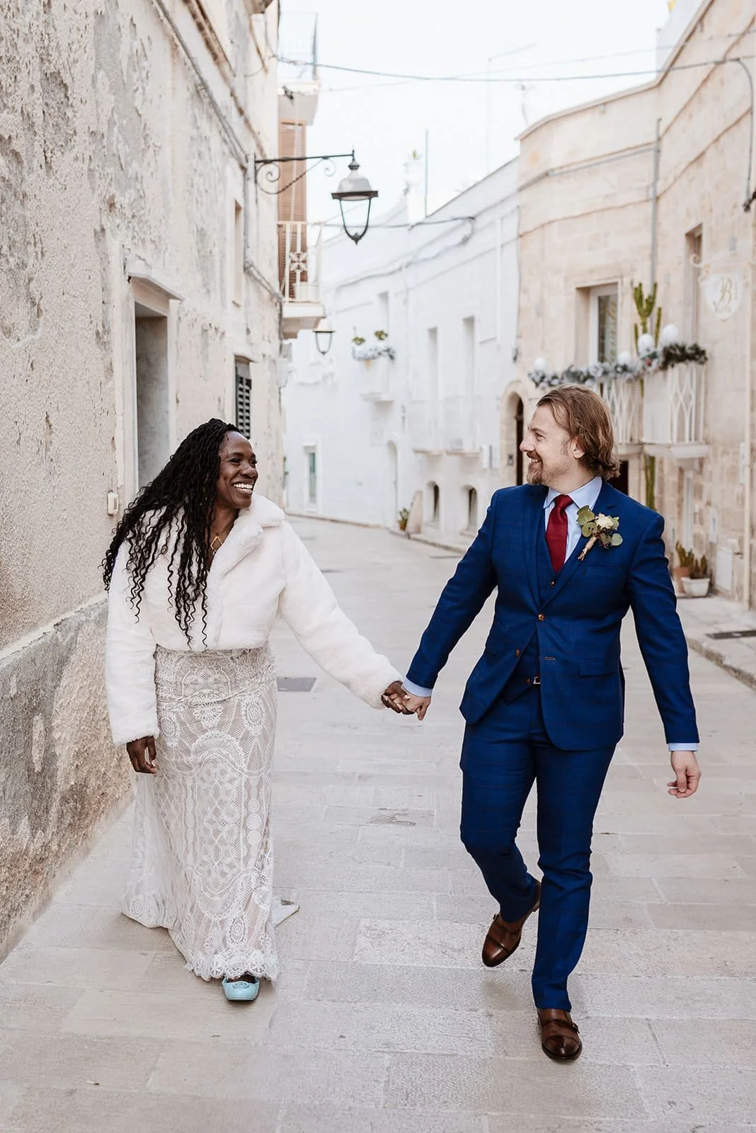 A happy couple holding hands, walking down a narrow street with white buildings, smiling at each other, dressed in wedding attire.