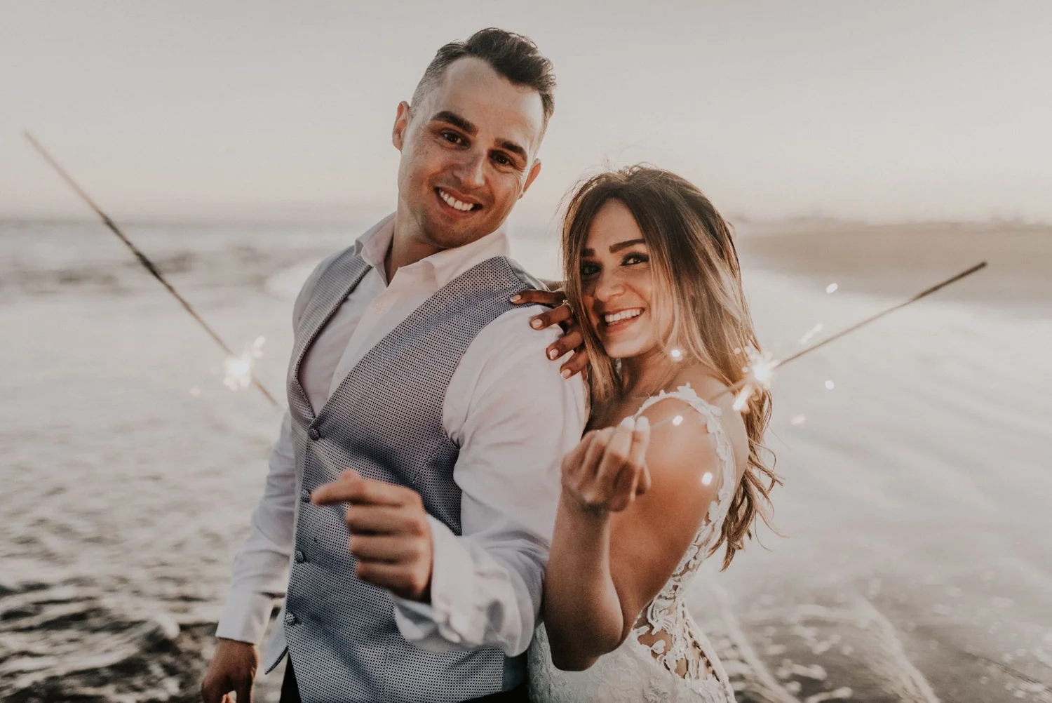 A happy couple embracing on the beach at sunset, holding sparklers, with the ocean in the background.