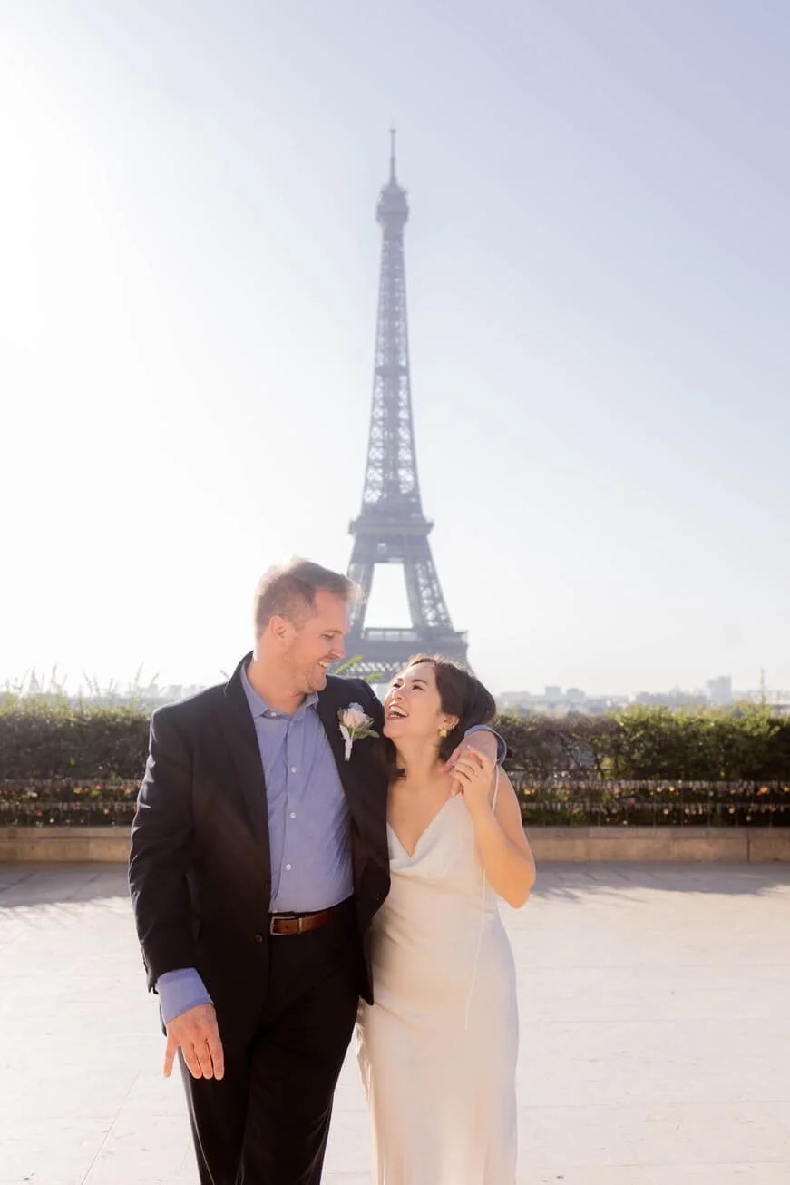 A couple in wedding attire smiling and laughing outdoors with the Eiffel Tower in the background.