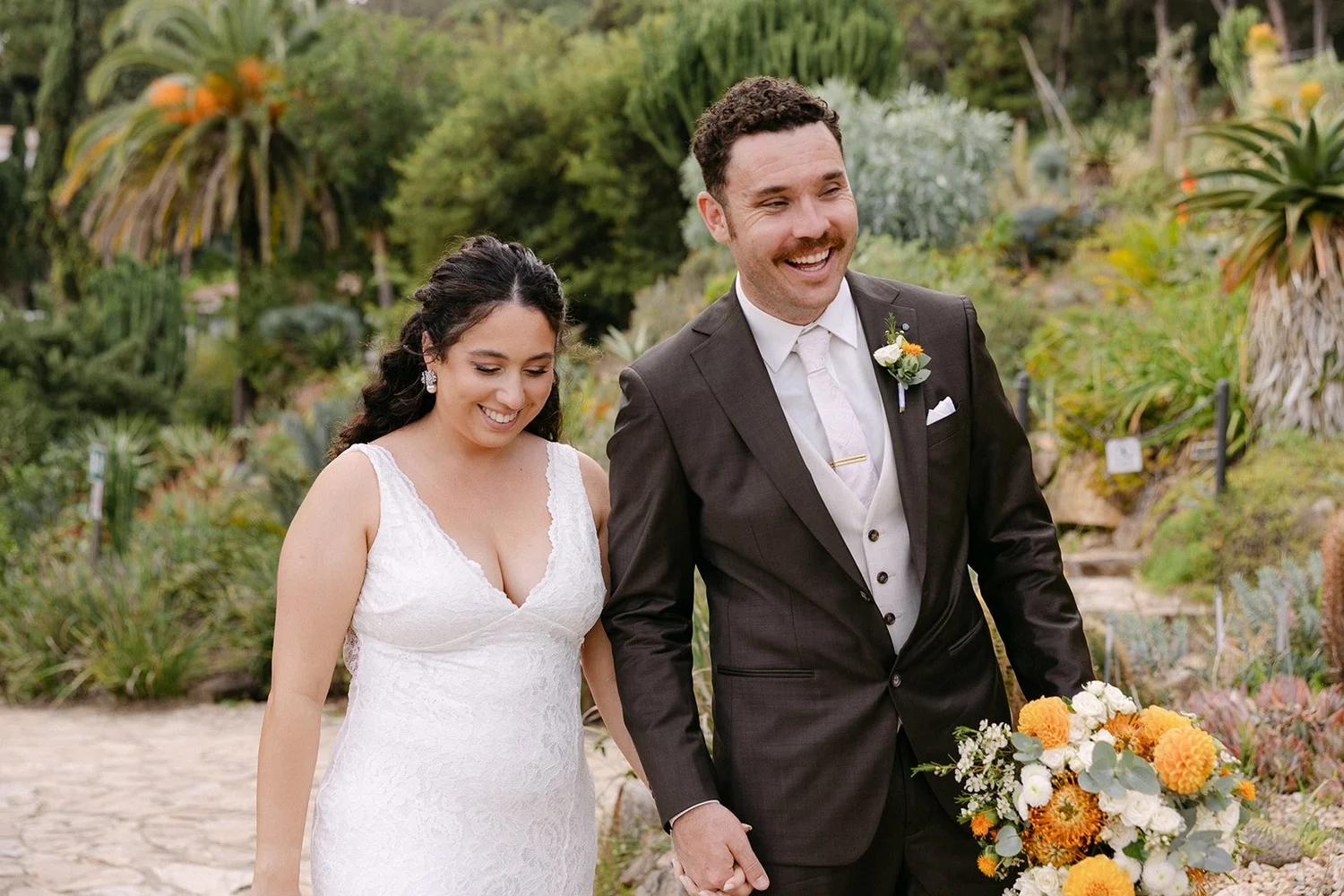 A bride and groom walking together outdoors, holding hands, with the bride wearing a white wedding dress and the groom in a dark suit, smiling.
