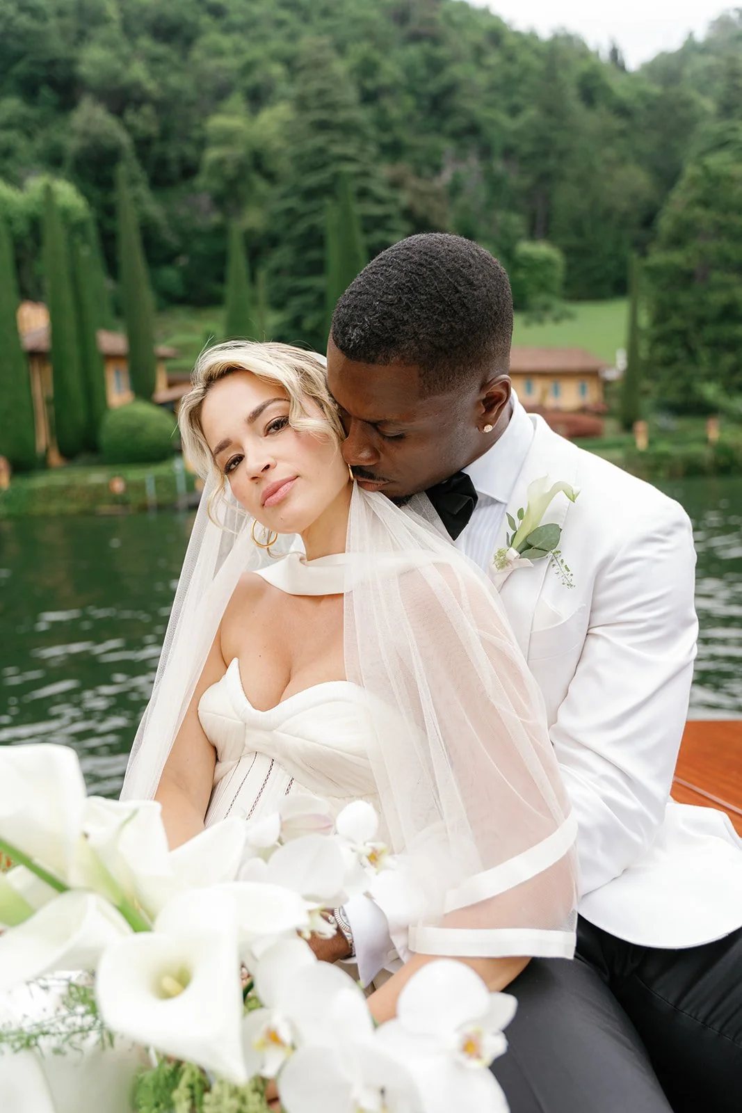 A bride and groom on a boat with a lake and greenery in the background, dressed in wedding attire, sharing an intimate moment.