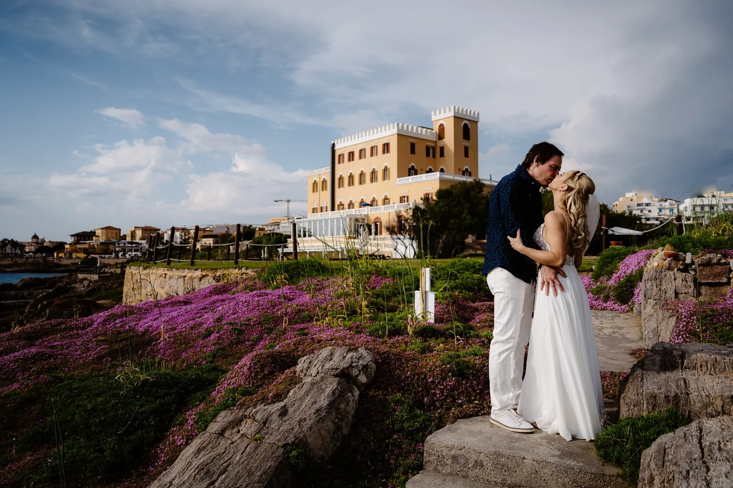A couple dressed in wedding attire sharing a kiss outdoors, with a large yellow building and a partly cloudy sky in the background, surrounded by purple flowers and rocks.