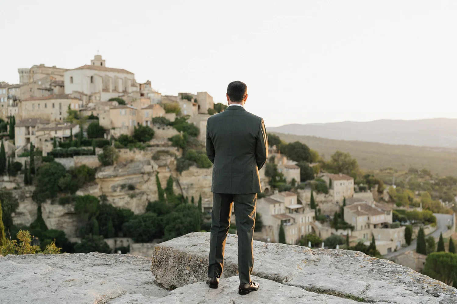 A man dressed in a suit standing on a rocky ledge, overlooking a hillside town with stone buildings and lush greenery at sunset.