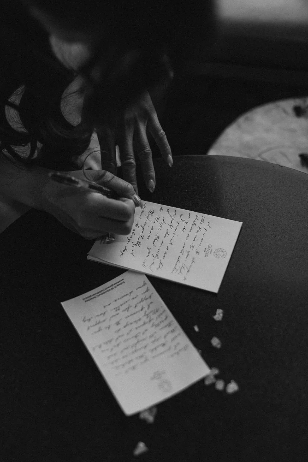 A person is writing a handwritten note on a piece of paper with a pen. There are additional papers and small heart-shaped objects on the dark surface beside the note.