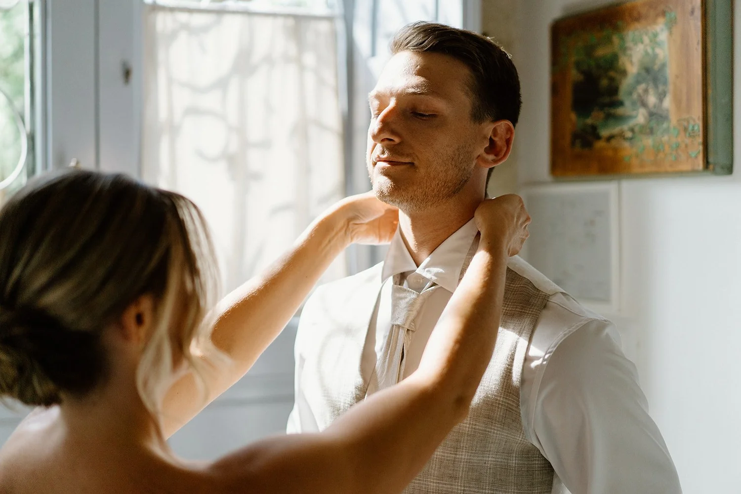 A woman helping a man adjust his tie in a well-lit room with a window and artwork on the wall.