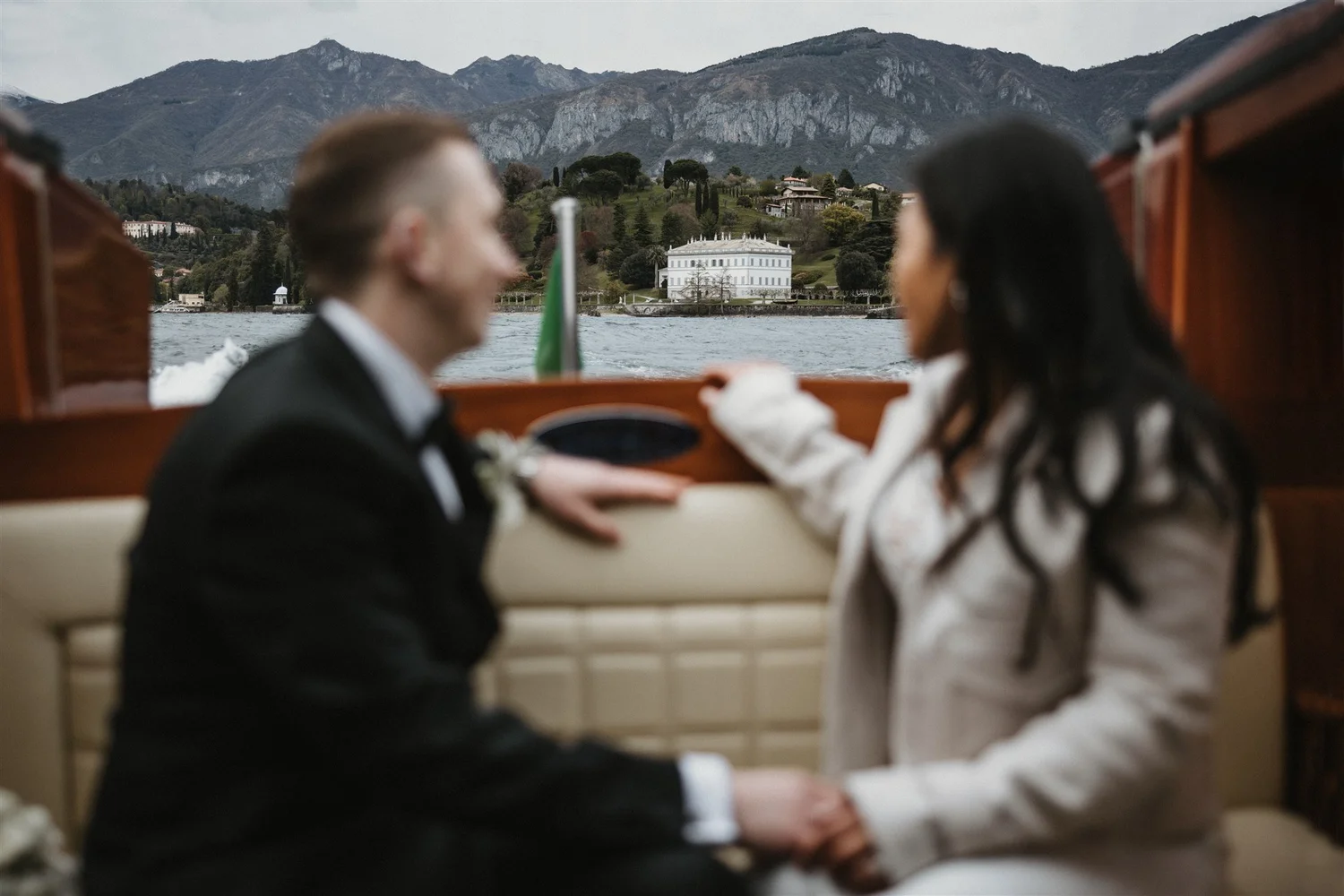 A couple dressed in wedding attire holding hands on a boat with scenic mountains and a large white house in the background.