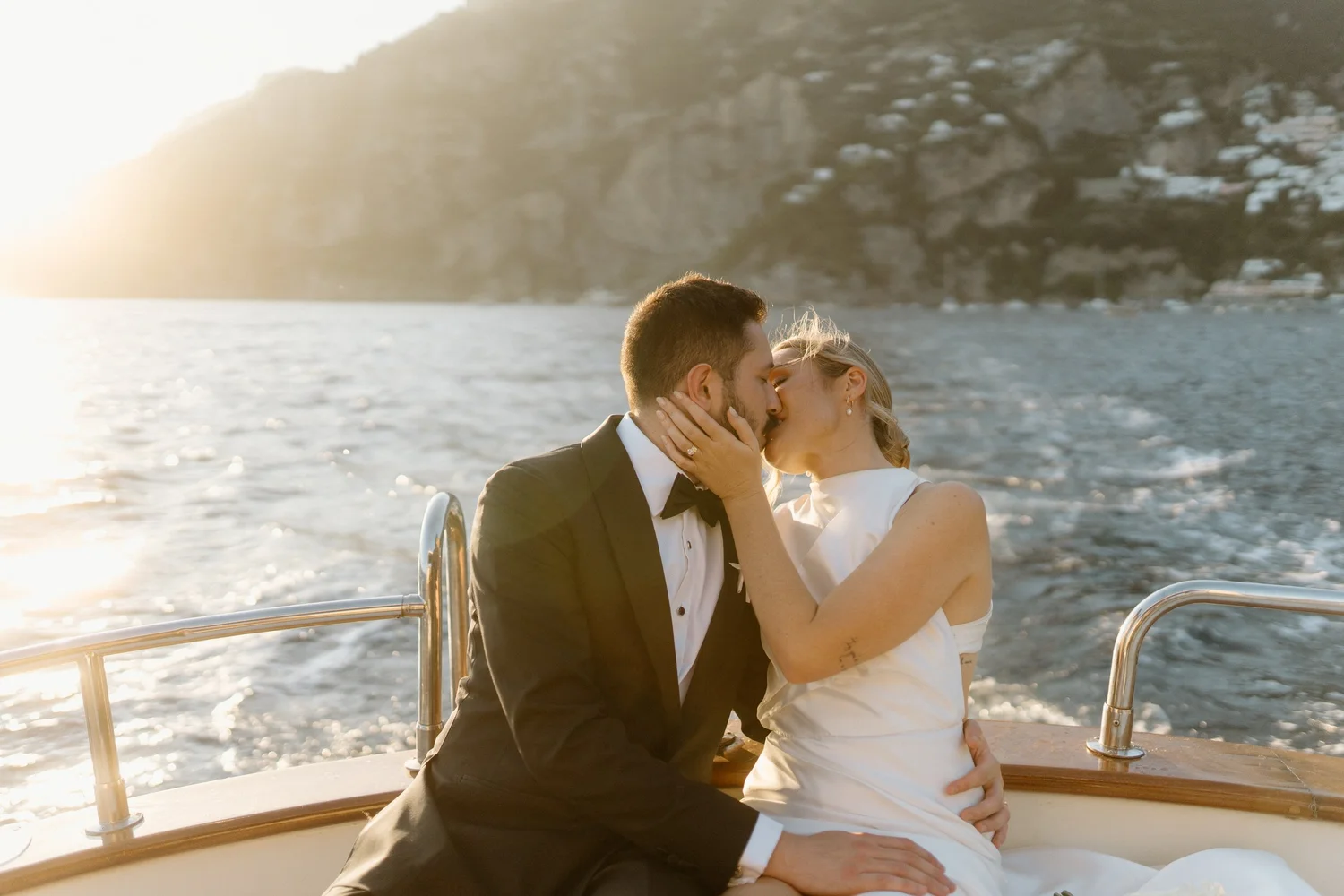 A couple dressed in wedding attire sharing a kiss on a boat with a scenic ocean and cliffs in the background during sunset.