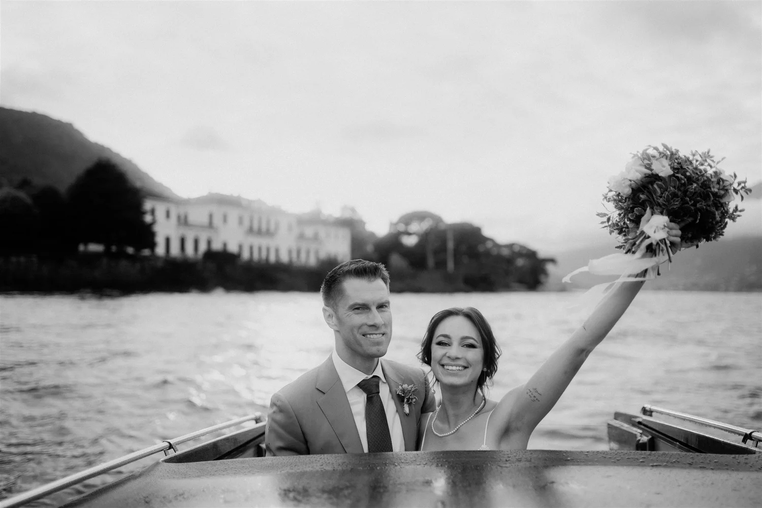 Black and white photo of a smiling couple on a boat, with a woman holding a bouquet of flowers, next to a water body with buildings and hills in the background.
