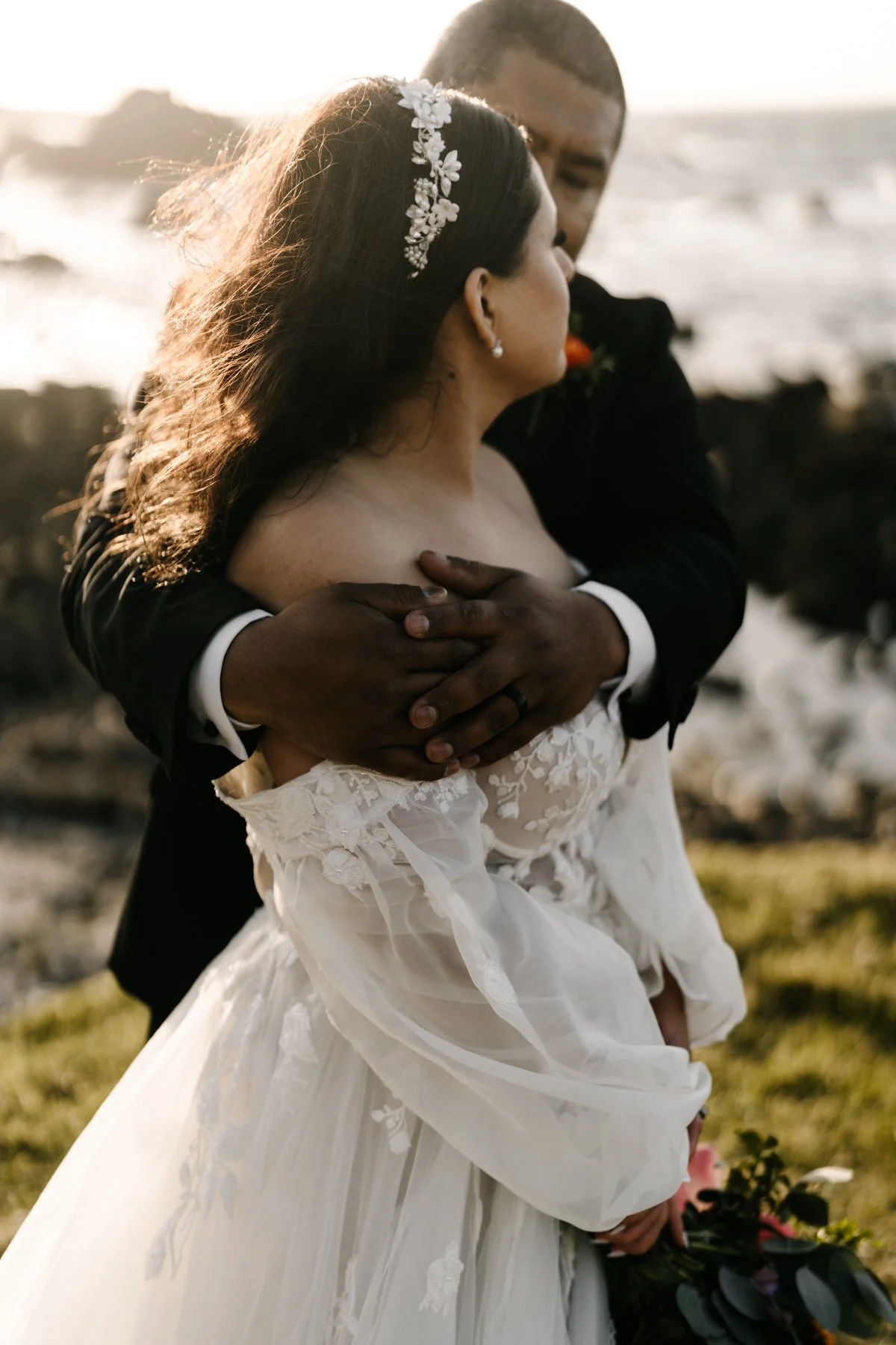 A bride and groom embrace on a beach during sunset, with the bride wearing a white lace wedding gown and floral headband, and the groom in a black suit, holding her gently.