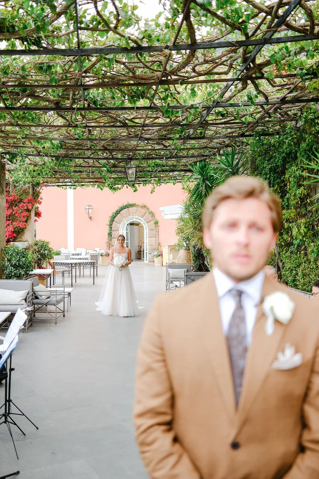 A wedding scene with a bride standing in the background holding a bouquet, and a groom in the foreground slightly out of focus. The setting is an outdoor courtyard with a pink wall, greenery, and decorative plants.