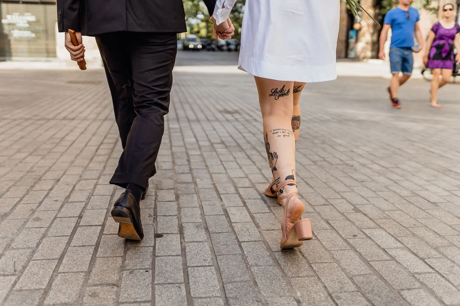Close-up of a couple walking hand in hand on a city sidewalk, focus on their lower bodies and legs, with a city street and pedestrians in the background.
